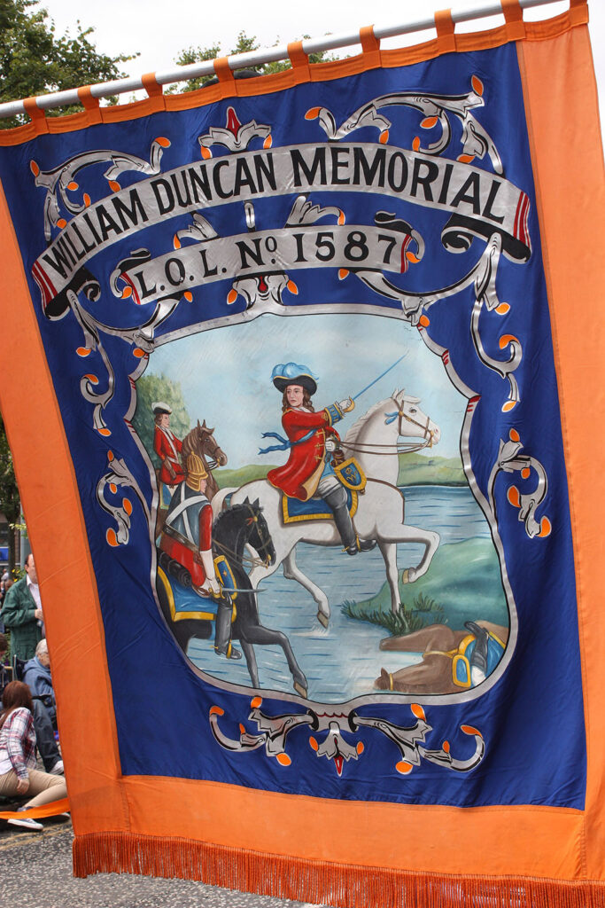 Oranje banier met een blauw middendeel met daarin de tekst "William Duncam Memorial, L.O.L. No. 1587" met daaronder de tekening van William III op zijn witte paard die zijn degen vooruitsteekt terwijl ze door een rivier lopen.