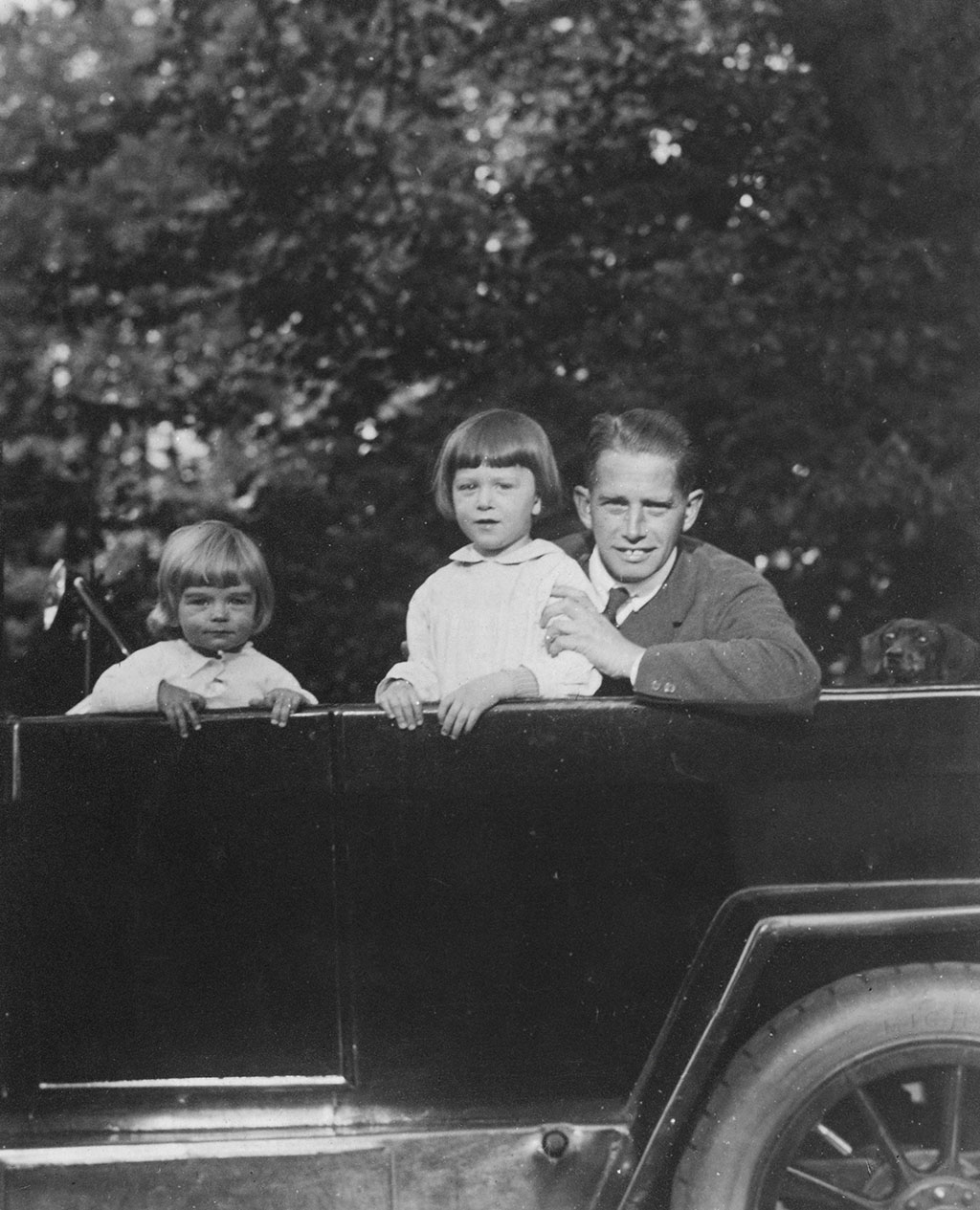 Zwartwit foto van het middendeel van een grote zwarte open auto. Rechts onderin zie je nog net een stukje van het achterwiel. In de auto zit een jonge man met een pak. Hij leunt over de zijkant en heeft zijn linkerhand op de bovenarm van een jongetje met een witte blouse. Hij staat in de auto en heeft zijn beide handen op de zijkant. Verder naar links staat een kleiner jongetje, ook met een witte blouse en ook met zijn handen op de rand van de auto. Achter de vader kijkt een teckel verschrikt omhoog.