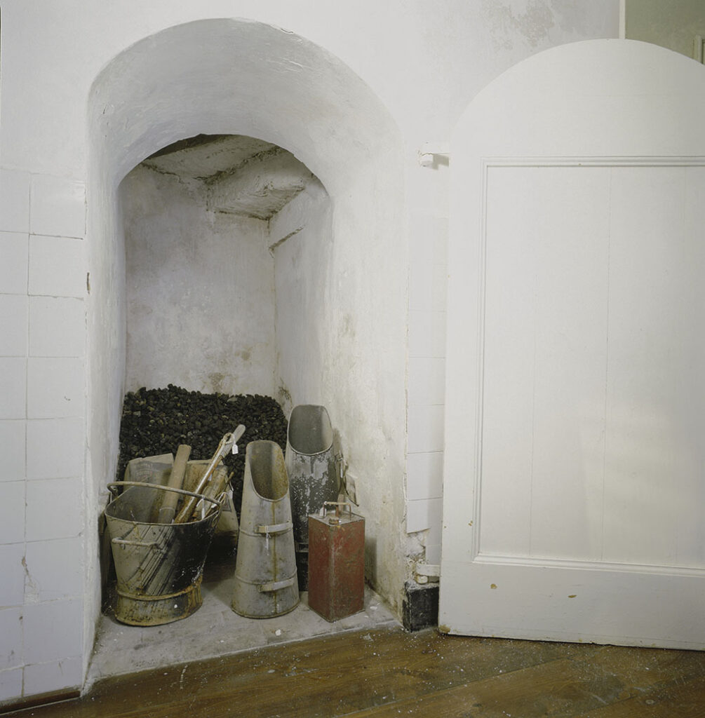 In een wand met witjes staat een deur open. Na een dikke muur zie je bovenin een nog een paar traptreden. Onder de trap liggen losse kolen, staan een paar kolenkits, een emmer met gereedschap en een petroleumblik.