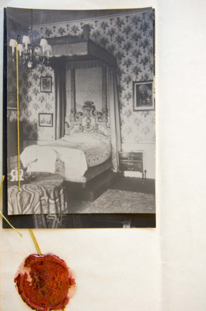 Een zwartwit foto van een kamer met een hemelbed op een witte pagina. Over de foto loopt een draad die vast zit met een zegel.