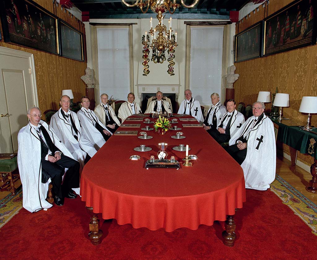 In een kamer staat een lange tafel met een rood tafelkleed. Aan de tafel zitten vooral oudere mannen die allemaal een witte cape dragen met een zwart kruis erop.