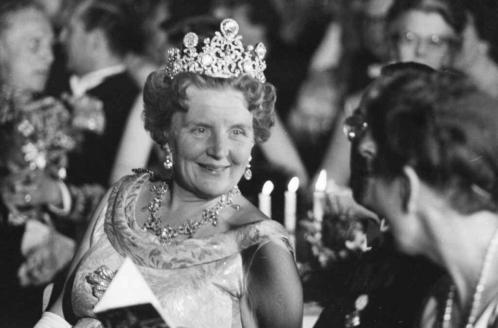 Zwartwitfoto van Koningin Juliana. Ze zit op een stoel, naast haar zien we nog net de bril van prins Bernhard. Ze praat met de dame die naast Bernhard zit. Ze glimlacht vriendelijk, draagt een grote, glimmende tiara met bovenop de Stuart diamant. Ze draagt parel oorbellen een eenvoudig collier en een jurk zonder mouwen.
