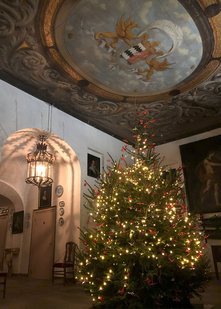 In de hal van het kasteel staat een grote kerstboom vol met rode gevouwen kraanvogels. Op het plafond de plafondschildering en op de achtergrond de boog naar de korte gang met schaduwen van een Venetiaanse lamp.