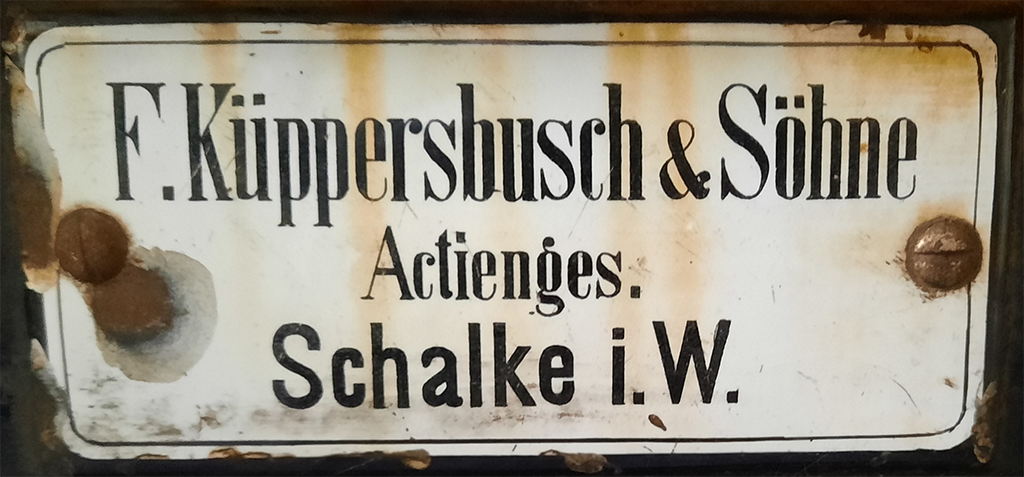 Geëmailleerd plaatje met daarop de tekst F.Küpperbusch & Söhne / Actienges. / Schalke i.W.
Het plaatje zit vast met twee schroeven links en rechts. Met name de schroef links is gaan roesten en roestplekken op het plaatje achtergelaten. Het witte email is op sommige plekken bruin geworden. Misschien door roest aan de achterkant. 