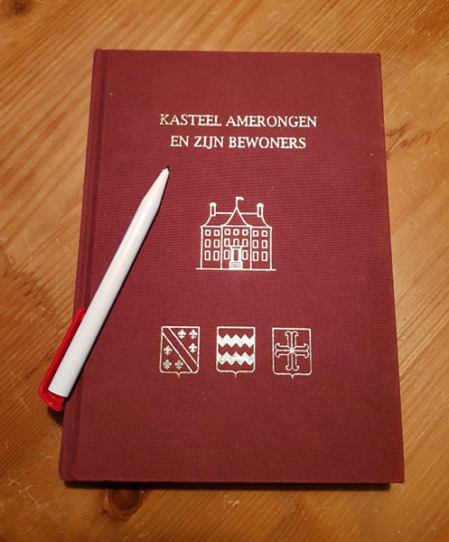 Rood boek met in gouden letters 'Kasteel Amerongen en zijn bewoners'. Daaronder een tekening van het kasteel en daaronder de wapens van de heerlijkheid Amerongen, de familie van Reede en de familie Bentinck. Op het boek ligt een witte pen.