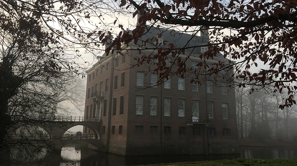 Een grauwe afbeelding van Kasteel Amerongen door de kale takken heen. Links de dubbele brug, het huis is zeven ramen breed en in het dak zitten twee dakkapelletjes. 