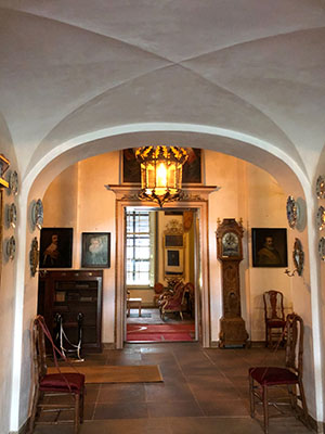 Doorkijkje door een korte gang, aan het eind van de gang hangt een grote lamp en zitten deuren naar de grote zaal. Links naast de deur een lage kast met een paar schilderijen erboven, rechts naast de deur een staand horloge. Door de linkerkant van de deur zien we nog net het uitzicht van een raam, door de rechterkant de muur. 