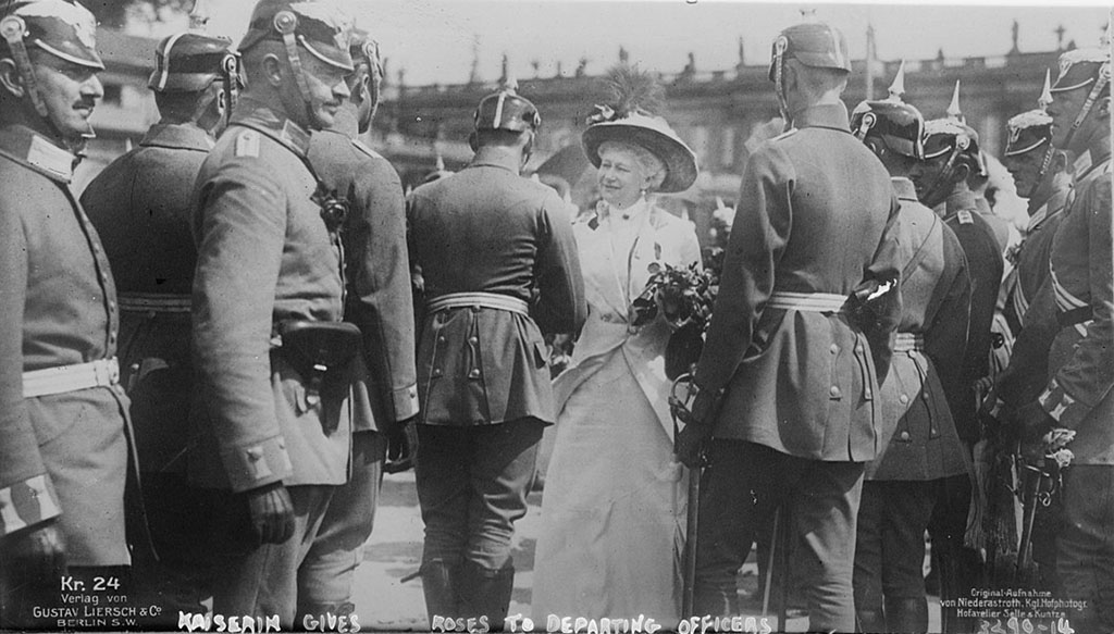 Tussen soldaten staat een elegant geklede vrouw met rozen in haar hand. 