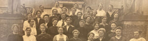 Groepsfoto van serieus kijkende dames, netjes maar eenvoudige gekleed.