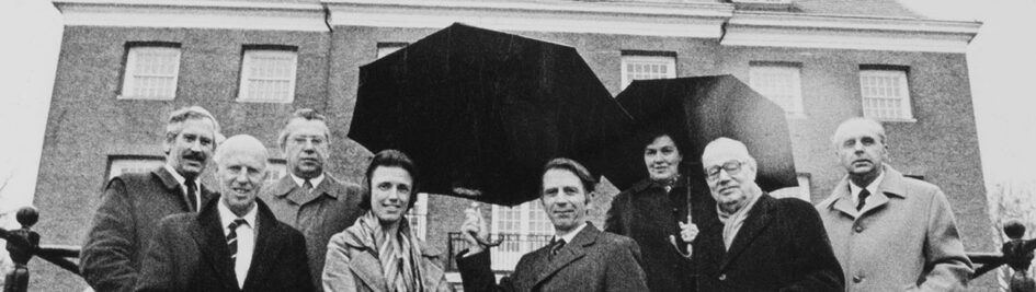 Zwartwit foto van mensen met jassen aan en paraplu's op voor het huis.