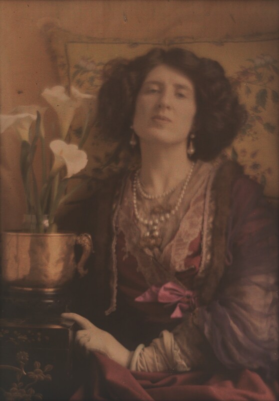 Een "half plate autochrome" foto, volledig in kleur maar wel met gedekte tinten. Op de foto een zittende dame die haar hoofd iets in haar nek heeft. Ze heeft een volle bos keurig gekapt haar. Ze draagt pareloorbellen en parelkettingen op een rode elegante jurk. Naast haar staat een bloeiende aronskelk in een koperen pot. 