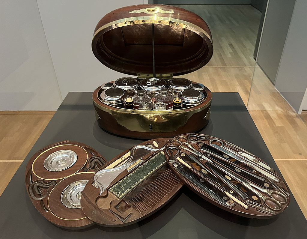 Een ovalen kist staat open en de verschillende lagen zijn tentoongesteld. In de kist zitten allerhande flesjes, potjes, haakjes, mesjes en ondefinieerbare voorwerpen.