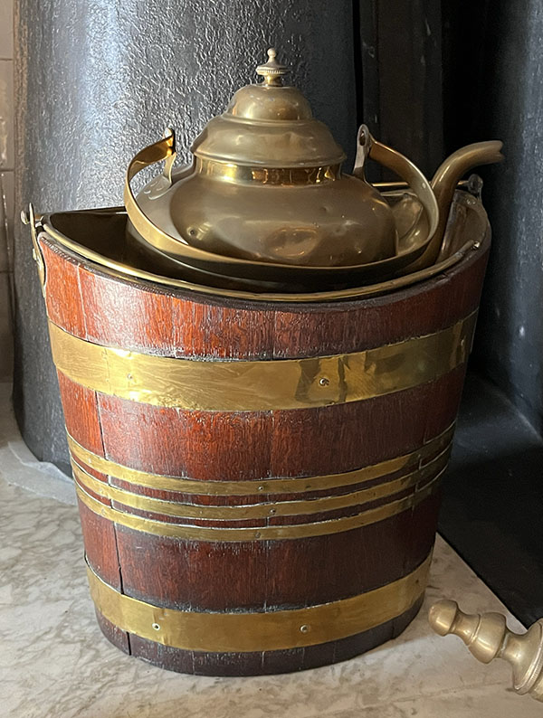 In een rode houten emmer met koperen banden staat een koperen ketel met een groot handvat.