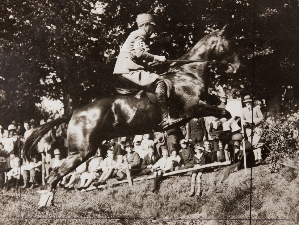 In een veld dat min omhoog loopt springt een paard met een militair op zijn rug. Op de achtergrond staan mensen te kijken. 