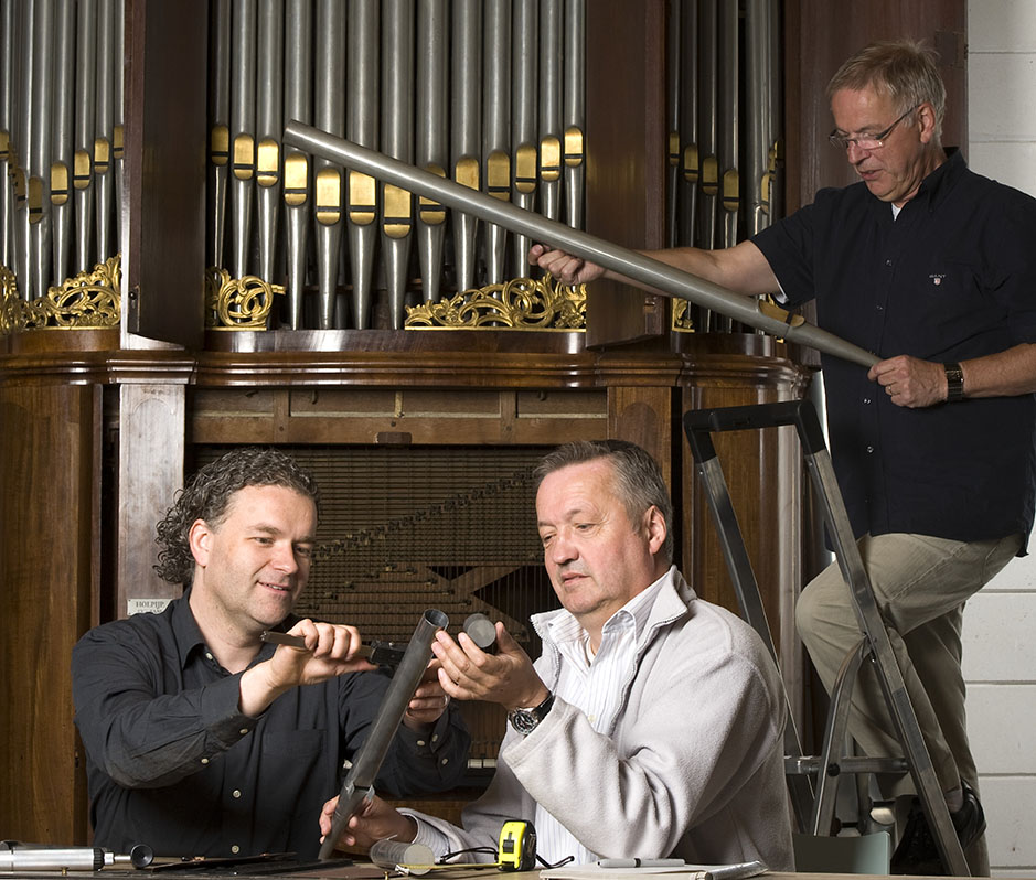 Voor een opengewerkt orgel staat een man op een trap met een orgelpijp in zijn handen. Voor het orgel zitten twee mannen, de ene meet de breedte van de orgelpijp die de andere man in zijn hand heeft.