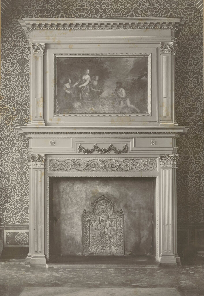 Zwartwitfoto van een schouw tegen een gedecoreerd behang. De schouw is eenvoudig gedecoreerd en heeft aan de bovenkant een schouwstuk. In de schouw staat een haardplaat, de vloer is kaal.