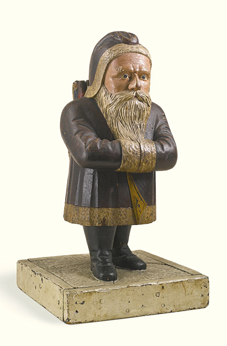 Een houten beeldje van een oude man met een bruine jas aan die is afgezet met bont. Op zijn hoofd een bruine muts met bont. Hij heeft een snor en een lange baard en heeft zijn handen in zijn mouwen gestoken.