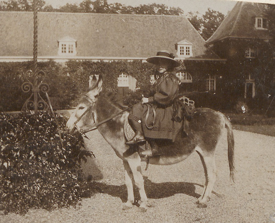 Op het voorplein van het kasteel zit een meisje in dameszit op een ezel. Ze draagt een lange jurk en een hoed met een brede rand.