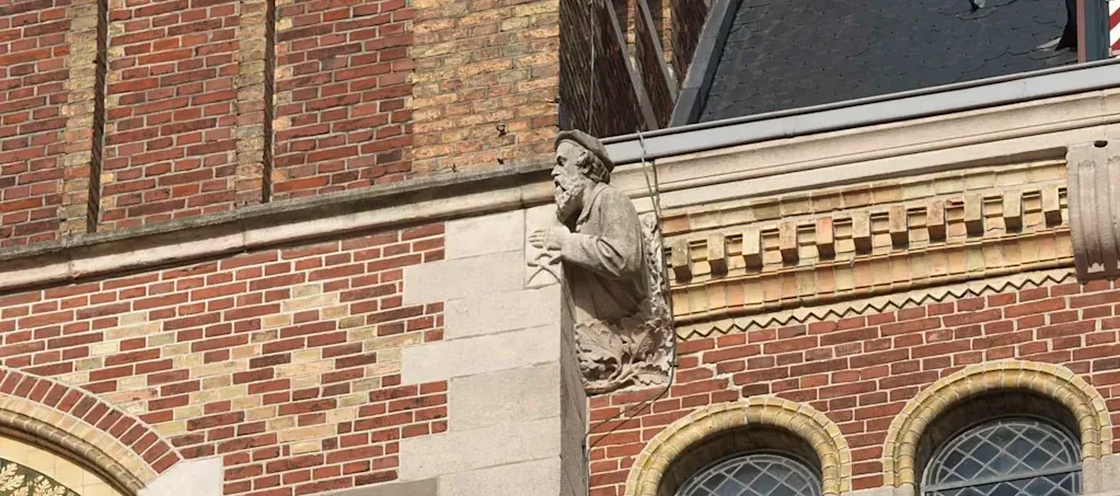 Op een gevel van rode baksteen met lichte ornamenten en natuurstenen lijst komt een beeldhouwde man om het hoekje kijken, hij draagt een plat hoofddeksel en heeft in zijn linker hand een stenenpakker. 