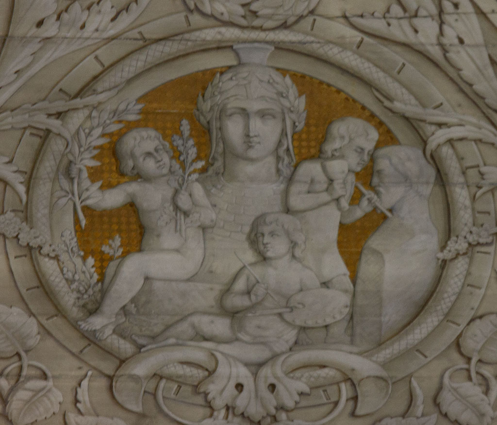 Medaillon met drie putti: één met lauwerbladeren, één met een schilders palet en één beeldhouwend aan een buste van een man met een baard. Op de achtergrond een vrouwenkop.