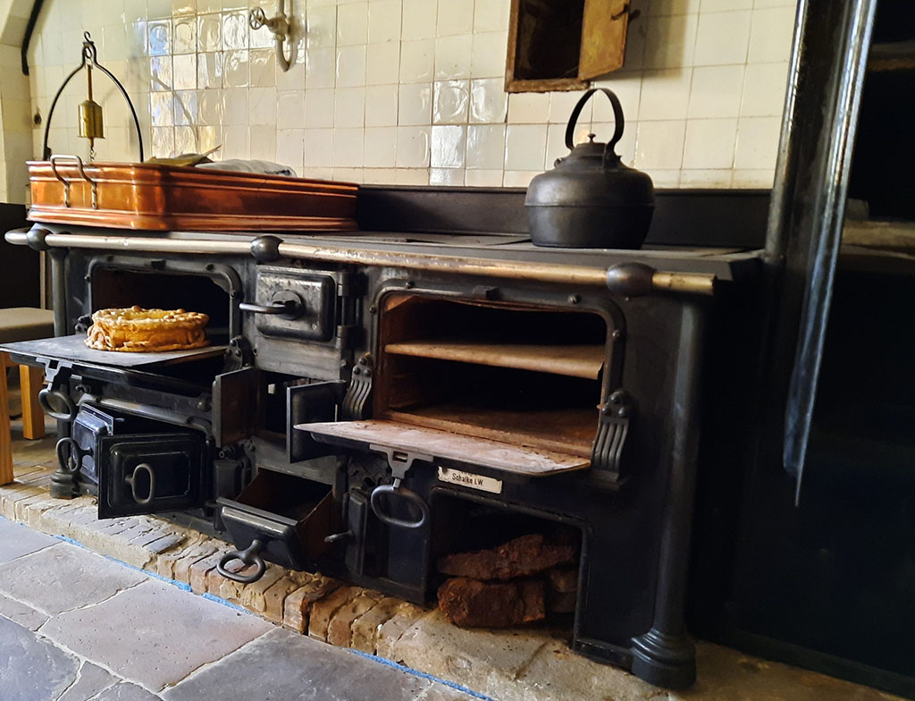 Een foto van een zwart gietijzeren fornuis schuin van voren. Alle kleppen van het fornuis staan open. In de oven links boven staat een taart. In een vak rechtsonder ligt turf. Op het fornuis staat links een grote rechthoekige koperen schaal en rechts een zwarte ketel. 