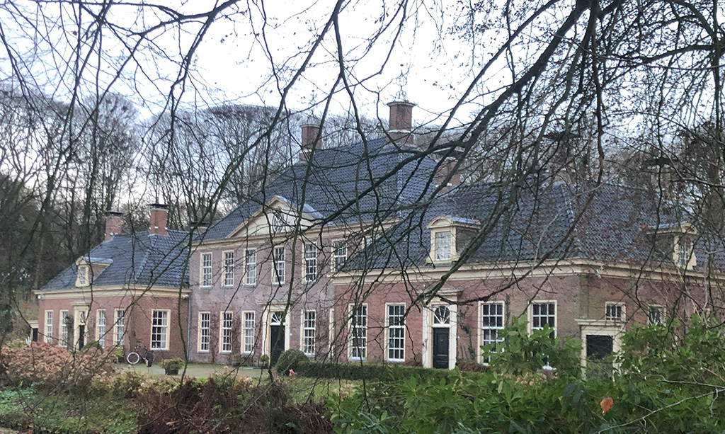 Foto van een twee verdiepingen hoog groot huis, de foto is door de kale bomen genomen. 
