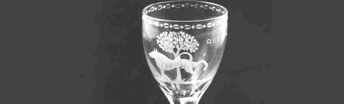 De kelk van een geslepen glas met afbeelding van een keeshond die zijn poot optilt bij een oranjeboom