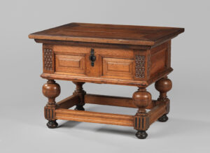 Zware houten tafel. de vier poten zijn met elkaar verbonden met latten, daarboven zitten sierlijke ronde vormen. Onder het tafelblad zit een grote kist, met daarop twee rechthoeken als versiering. De hoeken zijn geschubt. Voor op de 'onderlist' zit een zwart slot.