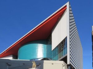 De gevel van Tivoli Vredenburg in Utrecht steek in een scherpe hoek af tegen de strakblauwe lucht. Rechts de gevel met de ronde raampjes, links een overhangend dak met een rode onderkant. Uit de glazen gevel, direct onder het dak, steekt een blauwgroene ronde vorm. Helemaal onderaan het beton van de oude concertzaal Vredenburg en daarvoor het beeld de verzekeringsengel van 'De Utrecht' (de 'Schele Maagd').