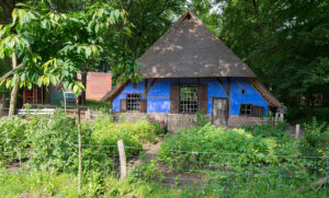 Blauw gestucte boerderij waar met balken (soort vakwerk). In het midden een groot raam met houten luiken en rechts daarvan een houten deur. Helemaal links een iets kleiner raam met luiken en rechts een klein raampje met luiken. De boerderij heeft een rieten dak. Ervoor een houten hekje en een moes- en kruidentuin. Achter de boerderij is bos.