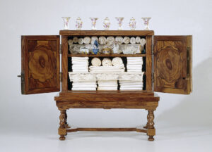 Bruin houten kast van marqueterie op een houten onderstel. De vier gedraaide poten zijn verbonden door een recht stuk hout die wigvormig uitlopen en zo de vier poten omvatten. De deuren van de kast staan open. De binnenkant van de deuren is gedecoreerd met gekleurd hout een groot bloemachtig motief. Op de kast staan om en om vier naar boven uitlopende vazen. Daartussen staan drie potten. Alle in meerdere lichte tinten gedecoreerd en van ongeveer gelijke grootte. De kast heeft twee planken. Beide planken hebben een kanten valletje. Op de bodem van de kast liggen drie stapel netjes opgevouwen wit linnengoed. Op de plank daarboven liggen aan de zijkanten twee stapels linnengoed, minder breed dan op de bodem. Tussen de twee stapels liggen twee brede stukken linnengoed met daarbovenop drie opgerolde stukken linnengoed. Op de bovenste plank is niet te zien wat er achter het valletje ligt. Ervoor staan porseleinen of aardewerken figuurtjes, erboven twaalf opgerolde stukken linnengoed.