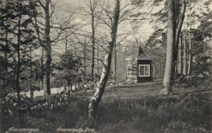 Een bos met van links naar het midden een pad. Rechts aan het eind van het pad staat een klein, donker huisje, een soort paviljoen, met een naar punt toelopend dak. We zien twee kanten van het huisje, met aan elke kant een groot vierkant raam met witte lijsten en een witte verticale lat in het midden. Op de voorgrond staan, vrij ver uit elkaar, twee bomen waarvan alleen de stam te zien is. Links op de foto staat een naaldboom met daarachter nog een boom en een struik. Meer naar achteren, rechts van het huisje staan nog meer bomen, evenals langs het pad ernaartoe.