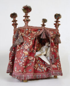 Pop, voorstellende de kraamvrouw, gekleed in een crèmekleurig satijnen jak, dat tot halverwege het bovenbeen reikt. Het jak is nauwsluitend en loopt vanaf het middel naar onderen toe uit. Onder het jak een enkellaags linnen hemd, daarboven een schoudermantel van kant.