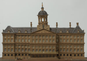 Een maquette van het paleis op de dam, een breed gebouw, 23 ramen breed. De gevel steekt bij de buitenste drie ramen wat uit en bij de binnenste zeven ramen. Boven de binnenste zeven ramen zit een fronton en met op de punt een beeld, op het dak in het midden een koepel. Het gebouw heeft een onderverdieping met tralies voor de ramen. Op de eerste verdieping een balkon in het midden. De eerste en de tweede verdieping hebben een combinatie van ramen met zes ruitjes en daar boven ramen met vier ruitjes, Tussen de ramen zitten pilasters.