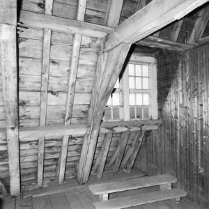 Zwartwitfoto van een zolder, duidelijk zichtbaar een dakspant en de planken van het dak. In het dak zit een raam en darvoor staat een trapje met twee treden