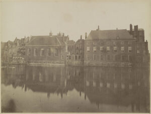Een zwartwitfoto van een vijver met aan de overkant allerhande gebouwen die aan elkaar zitten. Links een gebouw met vijf hoge boogramen en boven op het zadeldak een kleine klokkenstoel. Het grote gebouw rechts is zeven ramen breed, het heeft ramen in het souterrain, daar boven hoge ramen, daar boven iets lagere ramen en tot slot kleine ramen net onder de nok.