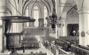 Zwartwit foto van het interieur van een kerk. Rechts tegen een pilaar de preekstoel, dan een kroonluchter. De kerkbanken staan eromheen gegroepeerd. We kijken door naar het koor van de kerk. Daar hangen rouwborden. Rechts in de zijbeuk een hogere kerkbank met daarachter een ruimte waar een gordijn voor hangt.