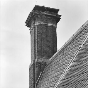 Zwartwit foto van een schoorsteen die hoog boven een leien dak uitsteekt. De bakstenen zijn afgewisseld met hardsteen. Bovenop zit een sierrand waar duidelijk stukken uit missen en een grote deksteen.