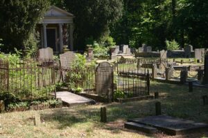 Foto van een oude begraafplaats in het groen. Het gras is groen, maar op sommige graven staat onkruid. De diversiteit aan stenen en hekjes is groot. Op de achtergrond tussen de bomen staat een mausoleum met zuilen en een timpaan.