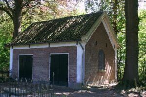 Een klein huisje met een puntdak in de schaduw van grote bomen. Aan de zijkant twee dubbele deuren, aan de kopse kant een raam met een boog.