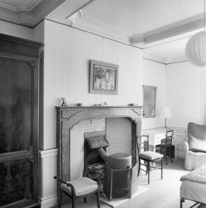 Zwartwitfoto van een kamer met een plafond onderverdeeld in vlakken. Een grote brede schouw met daarvoor een ronde kachel. Links naast de schouw een grote kast, recht een bureautje met daarop een schemerlamp en een spiegel aan de muur. Naast het bureautje een leunstoel. Op de voorgrond zien we nog net een klein stukje van een bed.