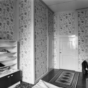 Zwartwitfoto van een kamer met een kattenbang. Links tegen de muur staat een kastje, rechts zien we nog net een stoel. Tegenover ons de deur en links daarvan een grote ingebouwde kast.