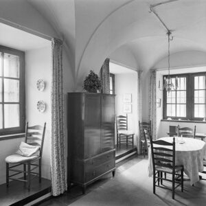 Een ruimte met een gewelfd plafond en drie ramen in diepe nissen. In de nissen staan stoelen en voor de nissen hangt een gordijn. Tussen de nissen links staat een eenvoudige afgesloten kast. In het midden staat een tafel met afgeronde hoeken en twee stoelen. Aan het plafond hangt een eenvoudige luchter.