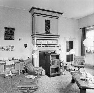 Zwartwit foto van een ruimte met een grote schouw waarvoor een stevige houtkachel staat. Voor de kachel staat een messing haardstel en twee leren leunstoelen, met ronde staande lampen. Links van de schouw staat een bureautje met daarboven wat foto's tegen de muur geplakt. Rechts van van de schouw een deur, rechts in de hoek een raam met plantjes op de vensterbank. Rechts voor een eiken koffietafel met een tegelblad. Op de tafel staan een koektrommel en wat snuisterijtjes. Bij de ene leunstoel ligt een breiwerk op de grond. De vloer is bedekt met gemeleerd tapijt.