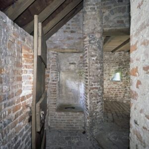 Een bakstenen doorsteek met recht voor ons een deur en daarachter een kleine ruimte met een gemetseld secreet met een houten plank met een gat erop. Rechts een gang met op de achtergrond een schietgat.