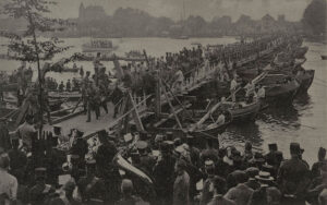 Zwartwit foto van de overtocht over de Schipbrug bij Dordrecht van het 1' Battillon, 15' Regiment Infanterie van het Korps Pontonniers tijdens de mobilisatie. Op de voorgrond speelt een militaire kapel. Op de achtergrond de stad.