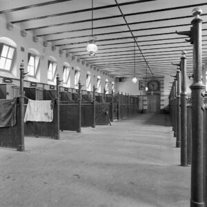 In een brede ruimte met hoge ramen zijn aan weerszijden stalruimtes afgebakend met schotten en tralies die vanaf de achterkant oplopen tot aan de muur. Op de achtergrond is een aantal boxen te zien en boven de deur in het midden is een hele grote klok verwerkt in de muur.