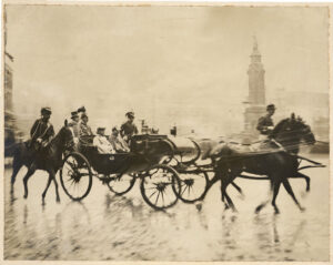 De keizer met koningin Wilhelmina en prins Hendrik in een open galarijtuig op de Dam. Op de achtergrond het 'Monument ter herinnering aan den volksgeest van 1830-1831', bekroond door het beeld van De Eendracht ('Naatje van de Dam') en het Commandantshuis.
