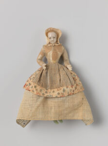 Een bleke pop van een vrouw met een strak lijfje, smalle taille en een wijde geruite rok met een geruit schort. Op haar hoofd draagt ze een kap.
