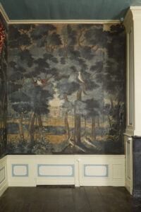 Een wand met een lage lambrisering met beige en lichtblauw, rechts zie je nog net de schouw. In het midden zit een deur, maar de beschildering loopt door over de deur. Aan de bovenste rand van de deur paleis soestdijk, op de voorgrond twee pauwen en een papegaai en een andere vogel in de bomen.